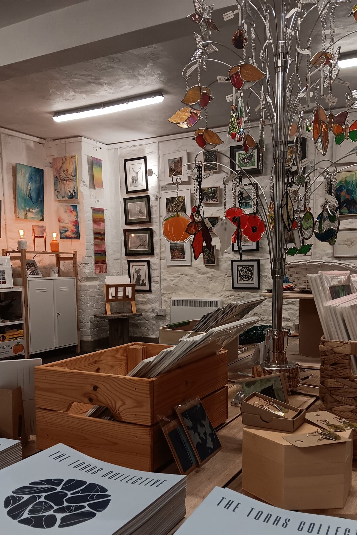 A retail display table in a gallery or studio space featuring stacks of 'The Torrs Collective' art booklets, wooden crates, and a decorative glass tree with colorful hanging glass ornaments.